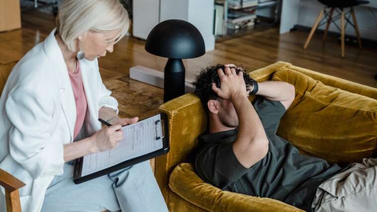 A woman takes notes while a distressed man sits on a yellow couch, holding his head in frustration.