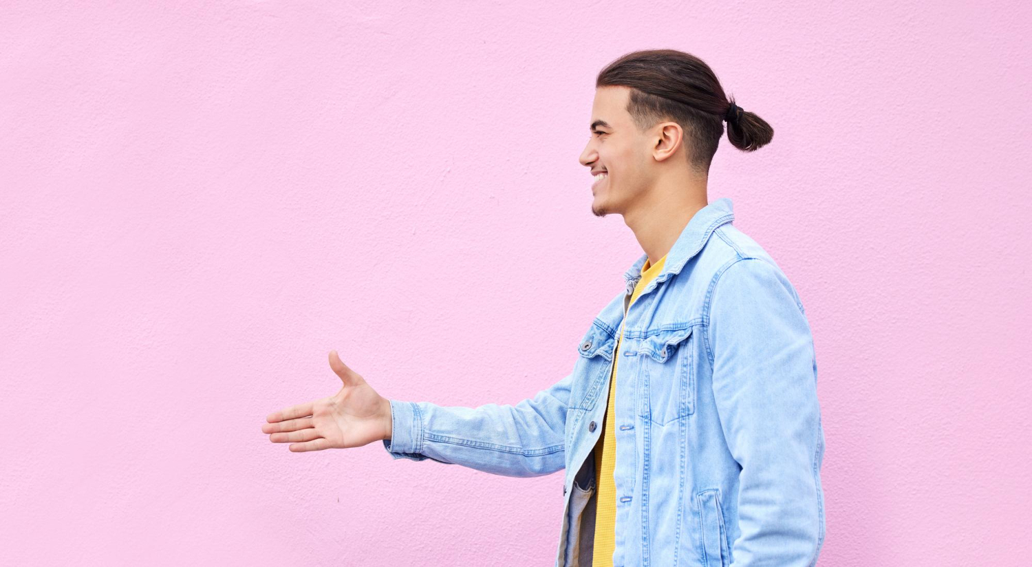 Young man in a denim jacket extends his hand for a handshake against a pink background.