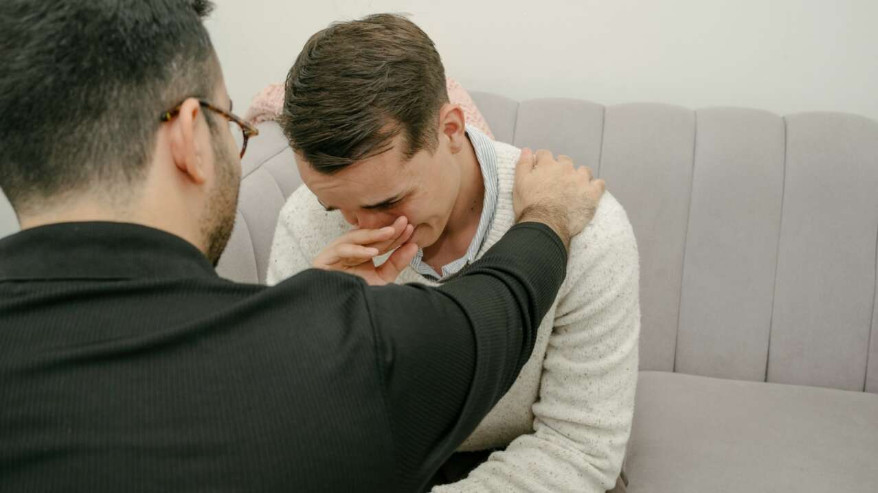 Two men, one comforting the other, sit on a light gray couch, expressing emotional support.