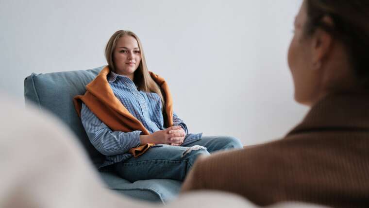 Young woman in a blue striped shirt and orange shawl sits on a couch, engaged in conversation.