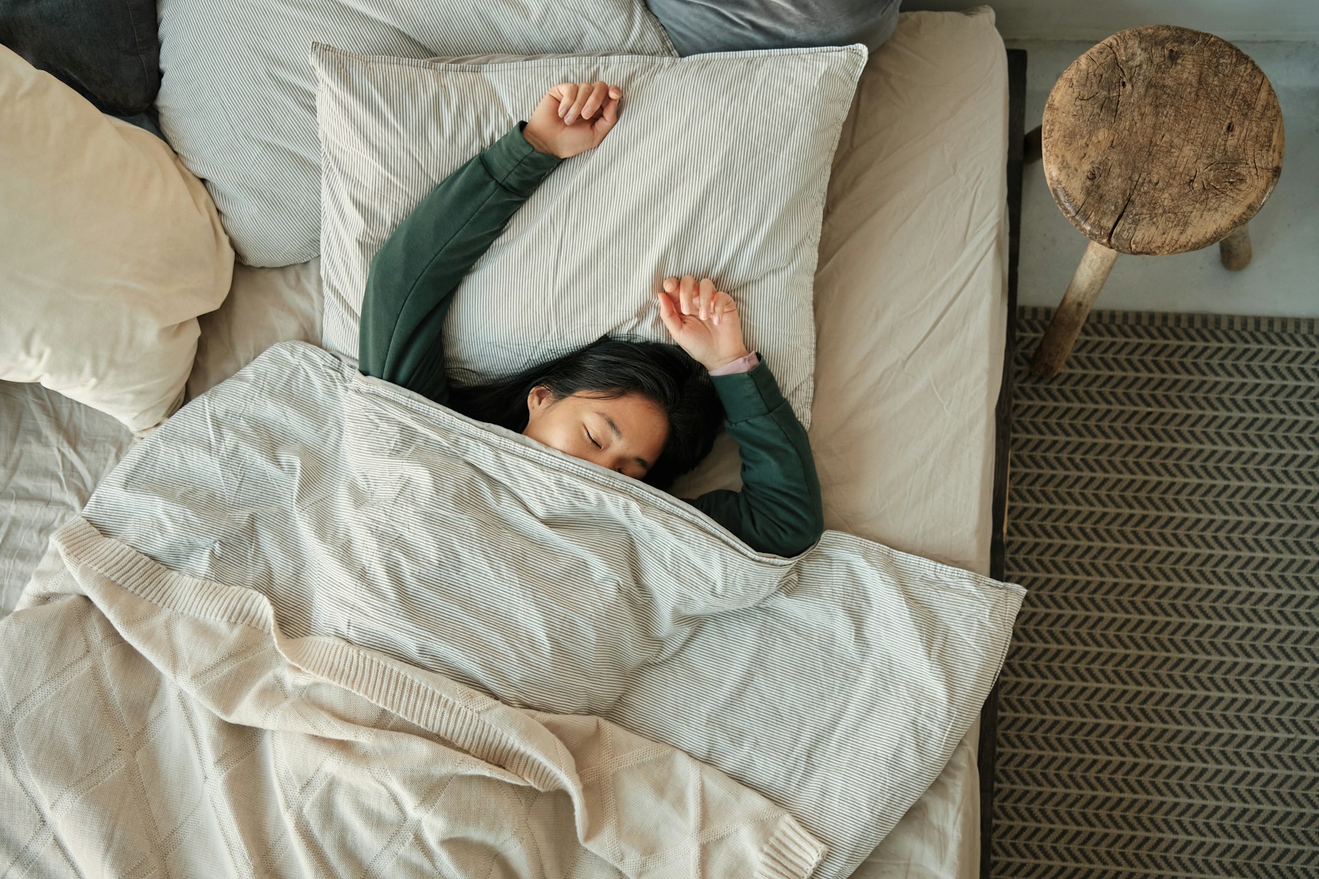 Young woman stretching in bed under a soft blanket, surrounded by pillows and a side table.