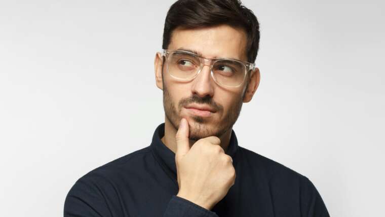 Man with clear glasses and dark shirt thoughtfully resting chin on hand against a light background.