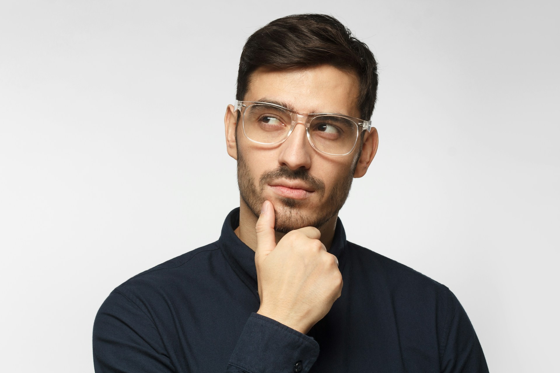 Man with clear glasses and dark shirt thoughtfully resting chin on hand against a light background.