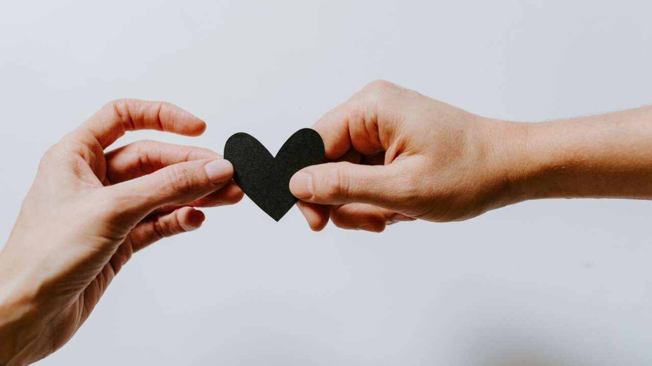 Two hands gently hold a black paper heart against a light background.