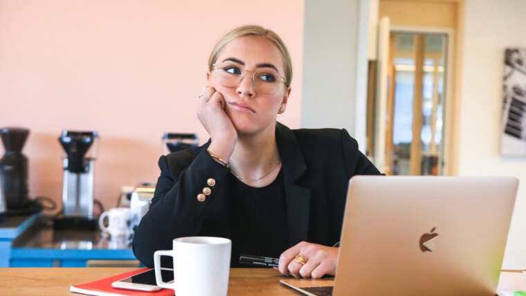 Woman with glasses resting her chin on hand, seated at a desk with a laptop and notebook.