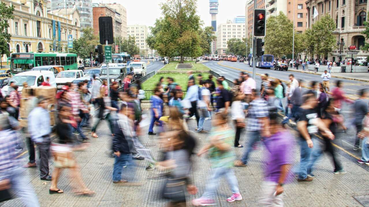Busy urban street with a crowd of people moving, cars, and a tall building in the background.
