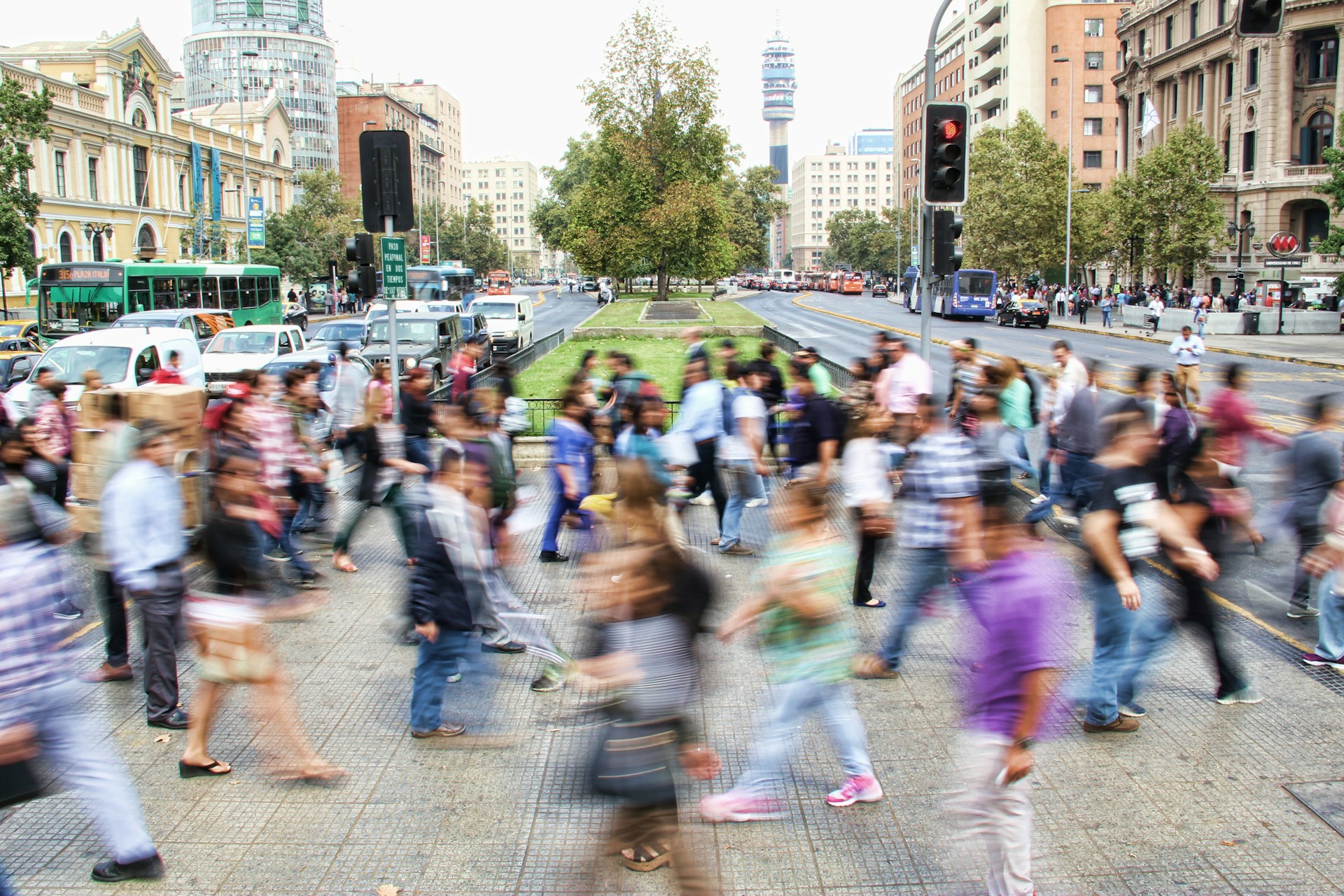 Busy urban street with a crowd of people moving, cars, and a tall building in the background.