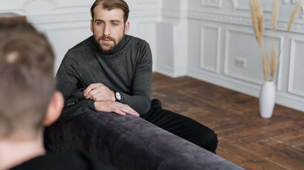 A man in a gray sweater holds glasses, sitting on a couch, engaged in conversation.