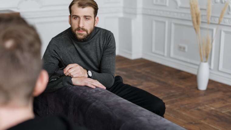 A man in a gray sweater holds glasses, sitting on a couch, engaged in conversation.