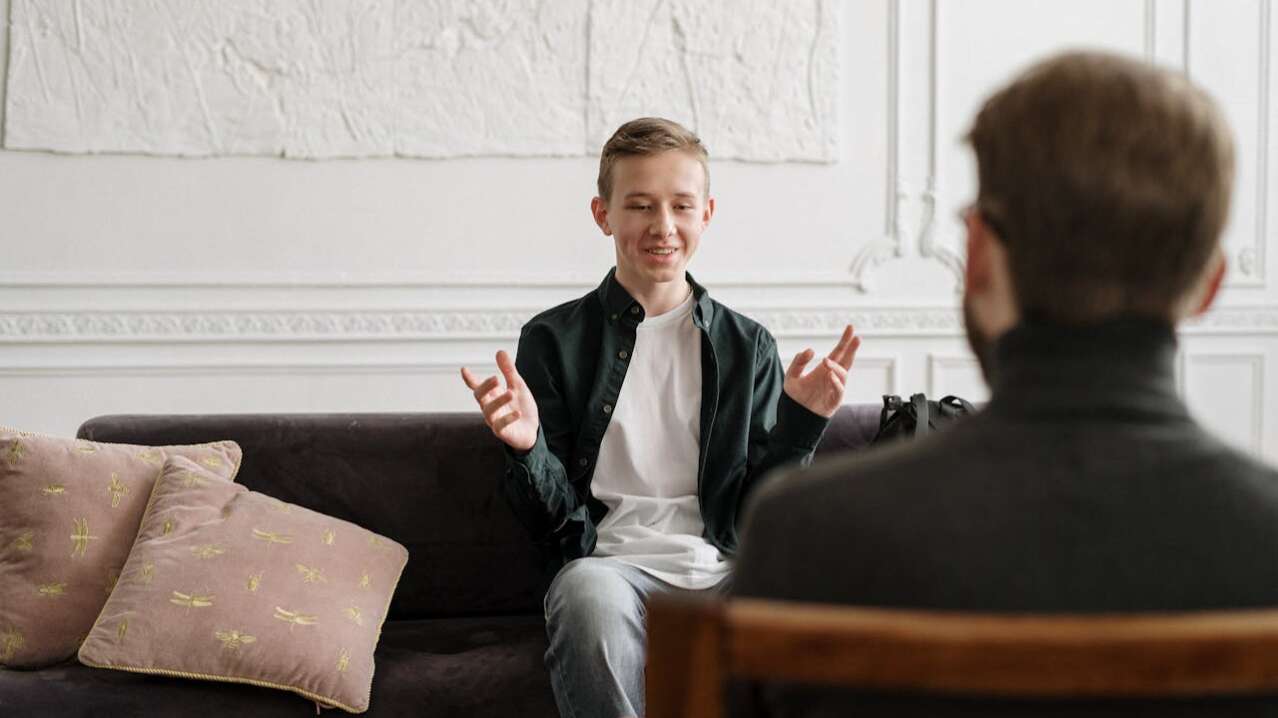 A young man in a green shirt smiles while gesturing during a conversation on a sofa.