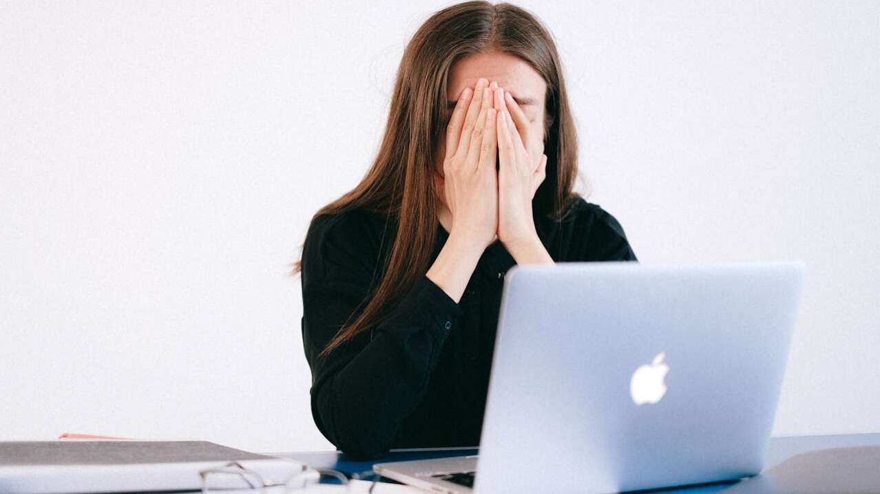 Stressed woman covering her face while sitting at a desk with a laptop and papers.
